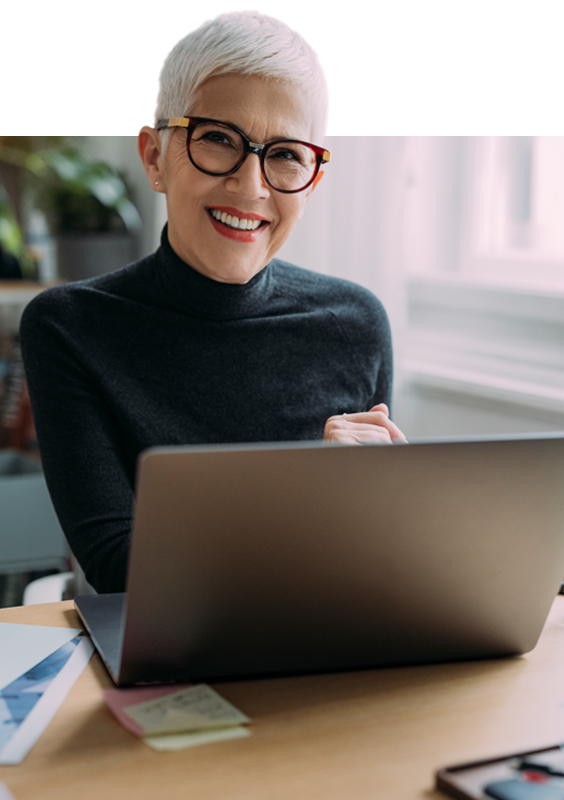 Seniorin arbeitet am Laptop im Büro Eine lächelnde Frau mit grazen Haaren und Brille sitzt am Schreibtisch und arbeitet an ihrem Laptop.